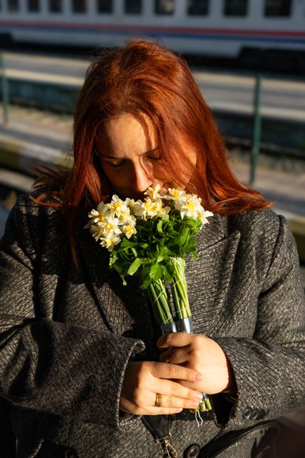 A woman with red hair, dressed in a dark grey coat, is holding a bouquet of fresh yellow and white daffodils with vibrant green stems and leaves. The bouquet is wrapped in clear plastic, highlighting 
