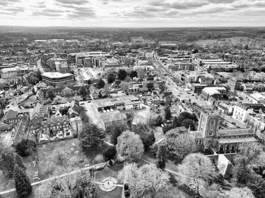 A black and white aerial view of Egham High Street showing a variety of buildings, streets, and clusters of trees, with a church featuring a tall spire in the foreground. The landscape extends to the 