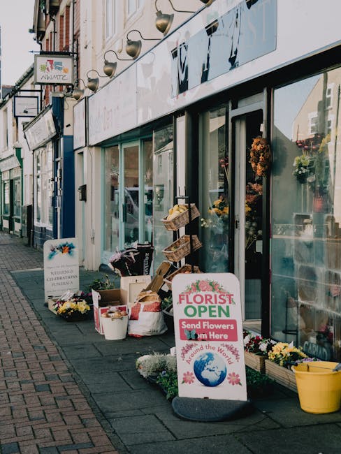 The storefront of a florist shop on Egham High Street featuring large glass windows displaying colorful floral arrangements inside. Outside, there are various buckets and baskets filled with fresh flo