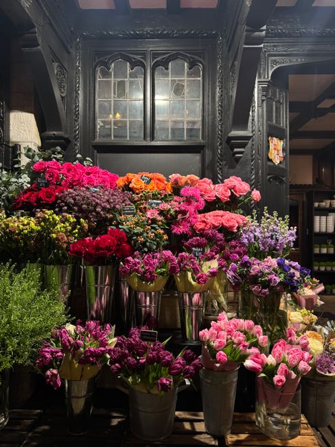 An indoor display of numerous flower arrangements and bouquets, predominantly featuring pink, purple, red, orange, and yellow blossoms, arranged in metallic vases and containers on a dark wooden surfa