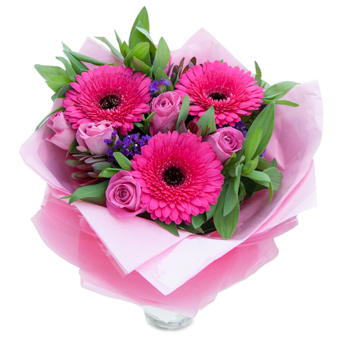 The charm of this bouquet lies in the contrast between bold and soft: three magenta Gerbera daisies burst open with exuberant petals circling dark, assertive centres, while surrounding roses in a muted lavender-pink curl inward with the reserved grace of a budding bloom. Little clusters of violet statice are tucked throughout like sprigs found on a riverside walk, adding speckles of cool colour and an airy, textural lift. A generous chorus of green foliage - broad, slightly glossy leaves that read as salal or eucalyptus - frames the blossoms, offering a crisp, almost dewy backdrop that heightens the pinks. The arrangement is wrapped with florist care: an inner veil of matte pale pink tissue cushions the stems, and an outer layer of translucent, slightly crinkled fuchsia-pink cellophane catches the light with a soft gleam. The clear glass vase's rim is visible at the base, a practical hint that the bouquet is intended for immediate display on a mantel or dining table. You can almost sense the scent: the gentle perfume of roses mingling with the fresh, subtle green of gerberas and foliage. I picture it brightening a home near the River Thames at Runnymede or a student flat beside Royal Holloway's tree-lined paths, carrying a message of affection, congratulations, or gratitude. Every element - from petal form to wrapping folds - reflects the steady hand and attention to detail you'd expect from a local Egham florist.